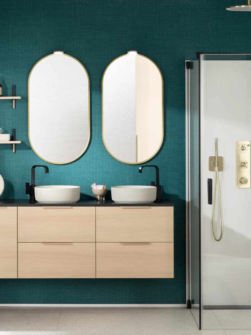 Badkamer met dubbele wastafel, afgeronde spiegels en zwarte kranen, smaragdgroene muur en glazen douchecabine met messing details in een moderne art-decostijl.